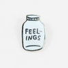 A small enamel lapel pin shaped like a bottle or jar with a white face, black outline, and the word "FEEL-INGS" in raised letters.