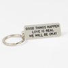 A silver metal keychain with a rectangular tag engraved with the text “GOOD THINGS HAPPEN / LOVE IS REAL / WE WILL BE OKAY.”