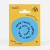 A blue spiral sticker on a yellow backing card with the text "GOOD THINGS HAPPEN. WE WILL BE OKAY. LOVE IS REAL." and smaller printed phrases including "It’s a sticker," "Makes me feel better," and "Hopeful but realistic."