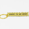 A gold-tone keychain with a jagged rectangular charm that reads "HAPPY TO BE HERE" in black letters.