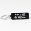 A black rectangular key tag with a metal split key ring, printed with the white text "HOME IS NOT A PLACE, BUT YOU ARE HOME".