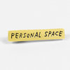 A rectangular gold-toned pin with the words "PERSONAL SPACE" in black hand-drawn lettering.