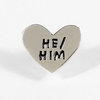 A silver heart-shaped pin with raised black text reading "HE/ HIM".