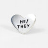 A heart-shaped metal pin with the black text "HE/THEY" embossed on the front.