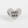 A silver heart-shaped pin with raised black text reading "SHE/HER".