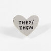 A silver heart-shaped pin with the black text "THEY/THEM" across the front.