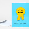 A blue greeting card with scalloped edges features a yellow chenille patch shaped like an award ribbon with the words "STILL ALIVE" and the printed word "CONGRATULATIONS" near the bottom.
