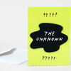 A neon yellow greeting card with scalloped edges, black question marks, and a black chenille-style patch reading "THE UNKNOWN" in white lettering.