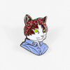 A cat-shaped enamel pin shows a white-and-brown cat bust wearing a red patterned headscarf and a light blue collared shirt with bright green eyes.