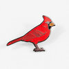 A red cardinal enamel pin with a black-outlined body, orange beak, gray crest, and pink feet.