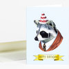 A folded greeting card shows an illustrated raccoon wearing a party hat and jacket above a banner that reads "HAPPY BIRTHDAY!".