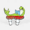 An enamel pin of a green frog reclining while holding a blue book, with red-and-white mushroom-like forms under its body and white stems below.