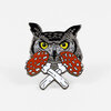 An enamel pin shaped like an owl face with bright yellow eyes, gray and black details, orange-red spotted wings, and crossed white bones beneath it.