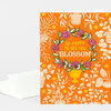 An orange greeting card with a floral wreath illustration and the text "SO HAPPY TO SEE YOU BLOSSOM," shown with a white envelope.