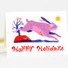 A folded greeting card shows a pink rabbit leaping beside a small tree and a sun, with the words "Happy Holidays" printed in red on the front.