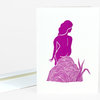 A folded greeting card with a magenta stylized figure standing among striped rock shapes and reed-like plants on a white cover.