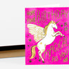 A bright pink greeting card features a white winged unicorn with a gold mane and horn and the text "HOPE YOUR BIRTHDAY IS AS MAGICAL AS YOU ARE" in gold lettering.