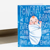 A blue greeting card with an illustration of a swaddled sleeping baby and hand-lettered text reading “CONGRATS ON YOUR BABY BOY. HE IS SO CUTE I CAN HARDLY TAKE IT. ANGEL ON THIS EARTH.”