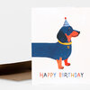 A folded birthday greeting card featuring a blue dachshund wearing a striped party hat and collar with the text "HAPPY BIRTHDAY" on the front.