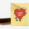 A folded greeting card with a metallic gold front, a large red heart with the words "I LOVE YOU" in white, and floral accents around the heart.
