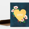 A folded greeting card with a large gold heart and the word "MOM" in white outlined letters, surrounded by white and orange flowers, green leaves, and small red berries on a dark blue background.