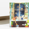 A folded greeting card shows two women in a bathtub reading, with plants, hanging lanterns, and a night sky visible through a window, and a blank envelope behind it.