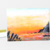 A greeting card with a watercolor-style sunset seascape featuring orange, pink, and yellow sky over water and dark rocky shoreline details.