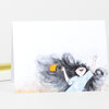 A folded greeting card shows an illustrated person with long dark hair, a light blue shirt, one arm raised, and an orange shoe or sandal in hand.