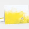 A folded notecard featuring a yellow-and-white illustration of a small figure standing in a field with arms outstretched and tiny scattered marks across the front.
