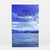 A paperback book titled "THE WRITER'S HANDBOOK" with the subtitle "A CONCISE SELF-HELP GUIDE FOR ESTABLISHED OR ASPIRING WRITERS" and the author name "DAVID SCHERER WATER" on a blue cover featuring water, mountains, and sky.