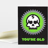 A folded greeting card with a black front featuring a white skull graphic inside green and gray starburst shapes and the text "YOU'RE OLD" in bright green.
