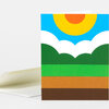 A folded greeting card with a stylized landscape design showing a blue sky, large yellow sun with orange center, white cloud shapes, green hills, and horizontal brown and green ground bands.