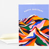 A folded greeting card with a sky-blue front featuring arched cream text that reads "HAPPY BIRTHDAY" above a colorful abstract mountain illustration.