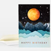 A folded greeting card with a starry night sky, a large orange moon, layered blue mountains, and the text "HAPPY BIRTHDAY" on the front, shown with a matching envelope.