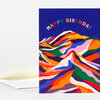 A folded greeting card with a navy front printed with arched multicolored "HAPPY BIRTHDAY" text above a layered abstract mountain design in bright rainbow colors.
