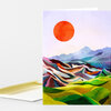 A folded greeting card with a colorful abstract landscape design featuring a large orange sun above layered multicolored hills and mountains.