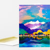 A folded greeting card featuring a colorful landscape illustration with a small house by a lake, layered mountains, trees, and a reflective water scene.
