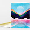 A folded greeting card with a colorful layered mountain-and-lake landscape illustration on the front and a blank white interior visible at the left edge.