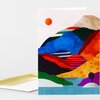 A folded greeting card with a colorful abstract landscape illustration featuring layered hills, a small orange sun, and a blank matching envelope partially visible behind it.