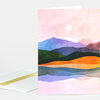 A folded greeting card featuring a pastel landscape illustration with layered colorful hills, a pale sun, and no visible text on the front.