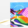A folded greeting card with an arched "HAPPY BIRTHDAY" message and a colorful abstract landscape of layered hills, dots, and stripes on the front.