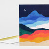 A folded greeting card with an abstract layered landscape of blue, green, red, orange, and yellow bands under a small crescent moon, with a white envelope behind it.