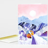 A folded greeting card featuring a stylized winter village scene with snowy mountains, purple hills, small houses, trees, and a pink sky with a white sun.