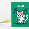 A folded greeting card with the word "HELLO!" and an illustration of a white cat wearing a red polka-dot hat riding a pink bicycle.