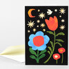 A folded greeting card with a black cover featuring colorful stylized flowers, stars, a crescent moon, and a white bird, with a blank white envelope behind it.