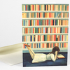 A folded greeting card shows an illustration of a woman lying on her stomach reading in front of colorful library bookshelves.