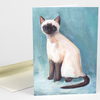 A folded greeting card features a painting of a Siamese cat with blue eyes, a dark face, ears, legs, and tail, and an off-white body against a blue background.