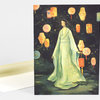 A greeting card featuring an illustrated woman in a pale green robe surrounded by hanging lanterns and a small crocodile at her feet.