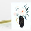 A folded greeting card with a watercolor-style floral illustration of pale peonies in a black vase with gold decorative details.