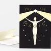 A folded greeting card with a black cover showing the words "YOU ARE MAGIC," a white star at the top, yellow radiating beams, and a white figure with outstretched arms surrounded by small stars.